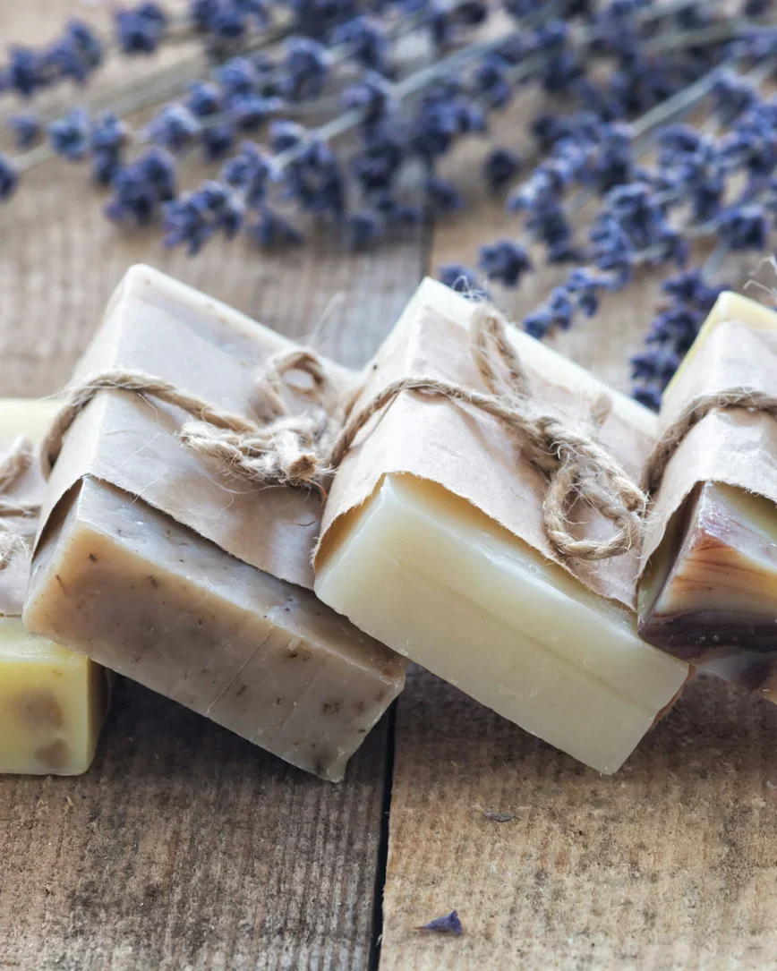 A flat-lay of neatly arranged grooming essentials: a bamboo toothbrush, a folded linen towel, a small ceramic soap dish with amber soap, and a sprig of eucalyptus on a warm alabaster background